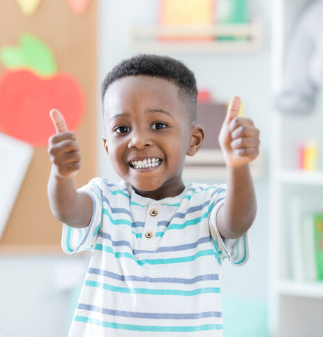 An adorable preschool age little boy smiles for the camera as he stands in his preschool classroom and gives a thumbs up.  He loves school!