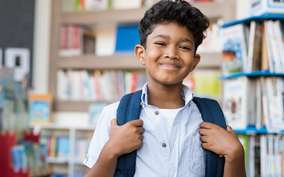 Portrait of smiling hispanic boy looking at camera. Young elementary schoolboy carrying backpack and standing in library at school. Cheerful middle eastern child standing with library background.